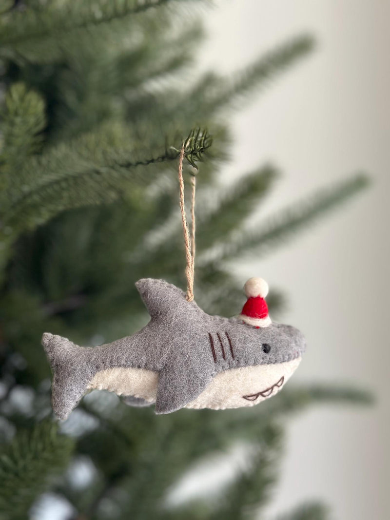 A delightful wool felt baby shark ornament is prominently displayed on a vibrant green Christmas tree. This gray and white shark features a festive red Santa hat with a white pompom. Its charming details include three stitched gills, prominent teeth, a black stitched eye, and a black stitched mouth. A jute string is attached, ready to hang this unique Christmas ornament.