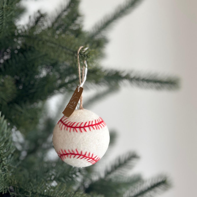 A handmade wool felt baseball ornament hangs on a Christmas tree, creating a festive, sporty atmosphere. The white ornament features red stitching, closely resembling a real baseball. It is suspended by a rustic jute string with a small tag, against a blurred green tree and white wall.