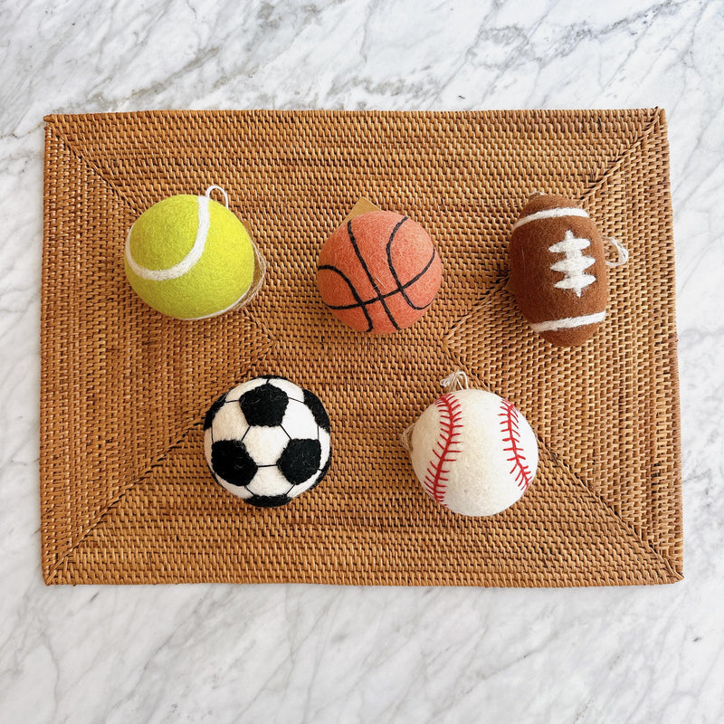 Five vibrant handmade felt ball ornaments, including a tennis ornament, a basketball ornament, a football ornament, a soccer ornament, and a baseball ornament, rest on a woven brown placemat, set against a clean white marble background. This wool felt ball ornament set features classic lines, accurately depicting each ball. Each one of them includes a jute string for hanging, making it a cheerful Christmas decoration.