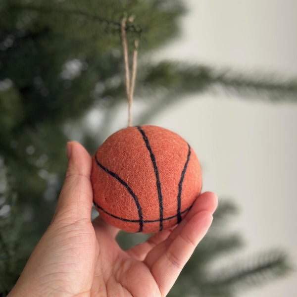 A handmade wool felt basketball ornament is gently held by a female hand, set against a softly blurred Christmas tree background. This round, orange ornament features black lines mimicking a real basketball's design and hangs from a natural jute string. It's a unique Christmas tree decoration that captures a sporty spirit.