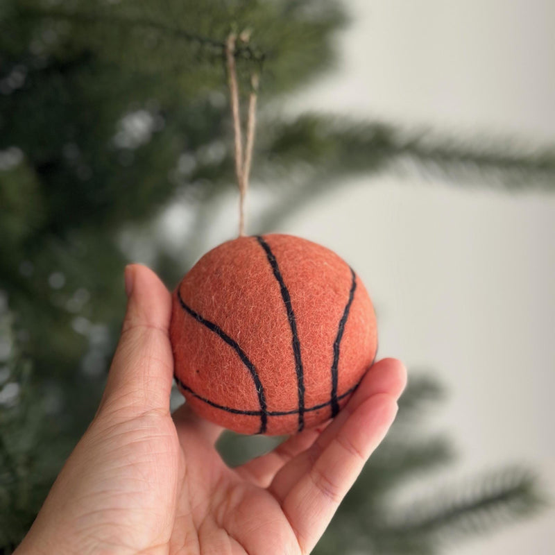 A handmade wool felt basketball ornament is gently held by a female hand, set against a softly blurred Christmas tree background. This round, orange ornament features black lines mimicking a real basketball's design and hangs from a natural jute string. It's a unique Christmas tree decoration that captures a sporty spirit.