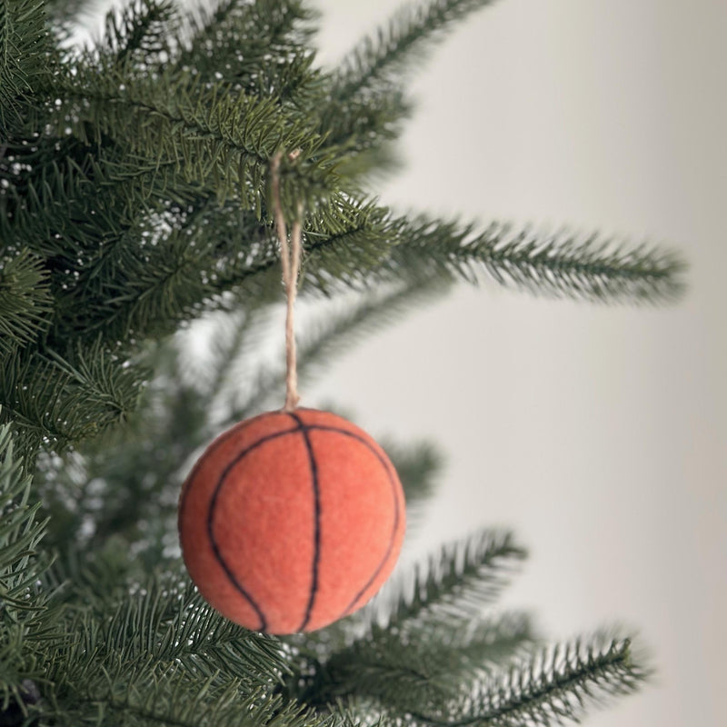A handmade wool felt basketball ornament hangs from the lush green branches of a Christmas tree, creating a festive, sporty atmosphere. The spherical orange ornament features realistic black stitched lines, mirroring a real basketball. A jute string allows it to dangle naturally, adding a handcrafted touch to holiday decor.