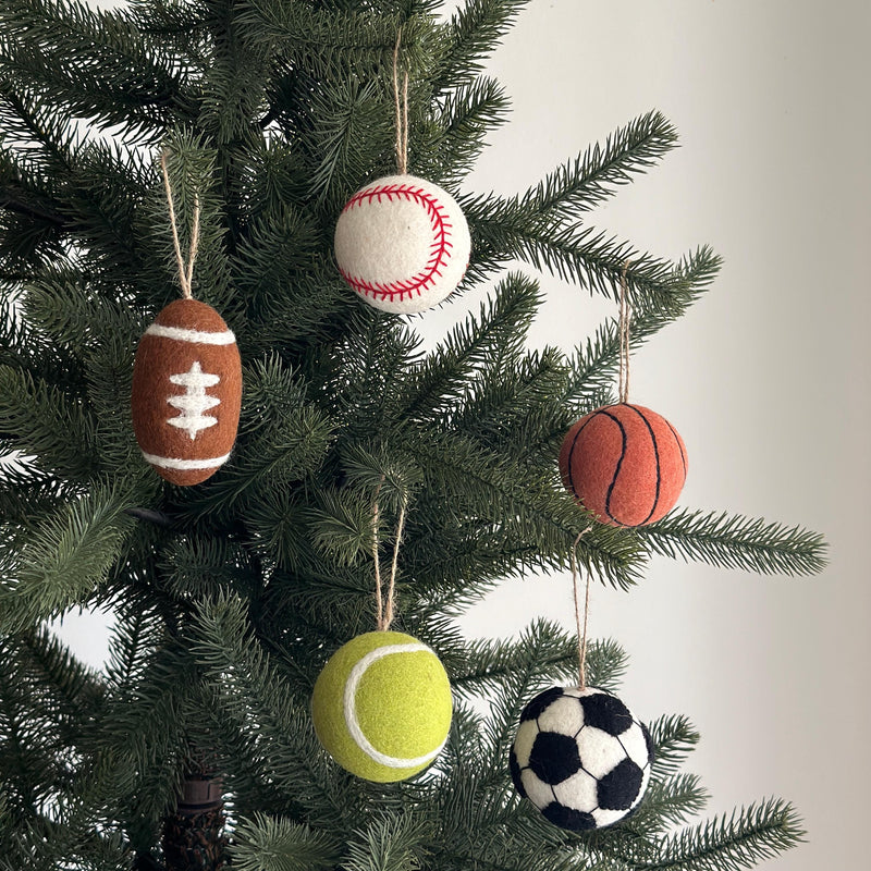 A set of six handmade wool felt ball ornaments hangs from a lush green Christmas tree. The ornament set includes a football ornament, a baseball ornament, a basketball ornament, a tennis ornament, and a soccer ornament. This unique sports Christmas decoration set is crafted from natural wool felt. Each one of the ornament set is suspended by a rustic jute string, adding a charming, handcrafted touch to the festive scene.