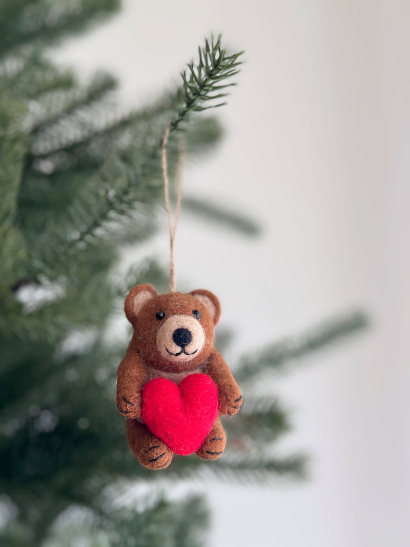 A charming felt bear holding heart ornament hangs from a pine branch, set against blurred green pine needles and a soft white wall. This light brown felt bear features small, rounded ears, black eyes, and stitched claws, holding a contrasting red heart in its paws. A jute string is attached for hanging, completing its cozy, handmade charm.