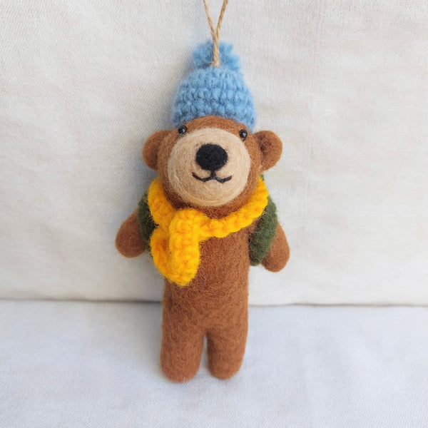 A handmade felt bear trekking ornament is displayed against a textured white backdrop. This charming brown felt bear wears a blue hat, a yellow scarf, and a green backpack, detailed with black eyes and nose. A jute string loop is attached to its hat for hanging.