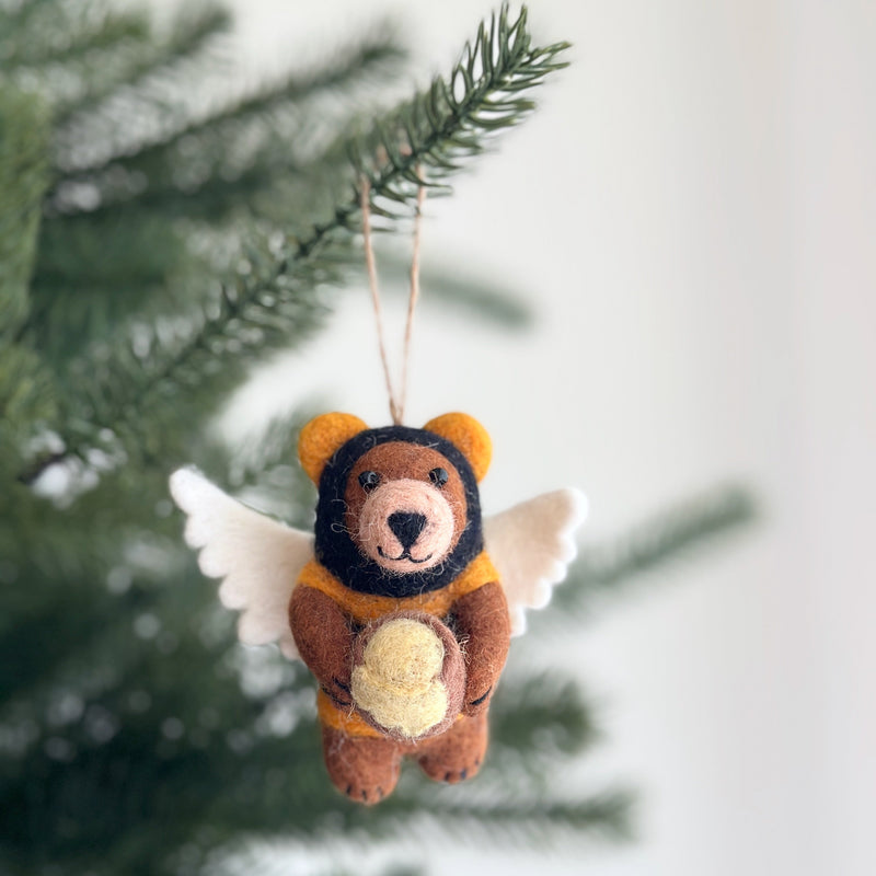 A felt bee bear ornament hangs gracefully by a jute string from a lush green Christmas tree branch, set against a soft white backdrop. This handmade felt ornament features a brown bear with fuzzy fur, small, round orange ears, and a black stitched nose. It wears a black and orange striped shirt with white felt wings on its back and clutches a tiny honey jar.