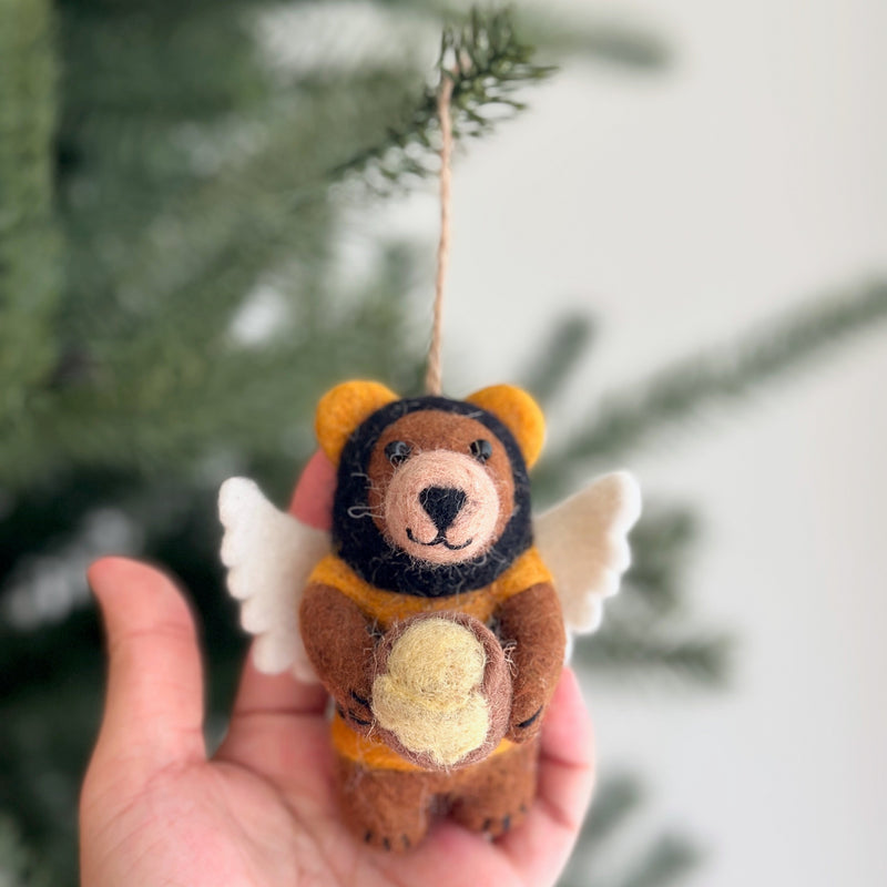 A charming felt bee bear ornament is held by a female hand, suspended by a jute string against a blurred Christmas tree backdrop. The brown felt bear ornament is dressed in a yellow and black bumble bee costume with white felt wings and holds a small beige honey jar.
