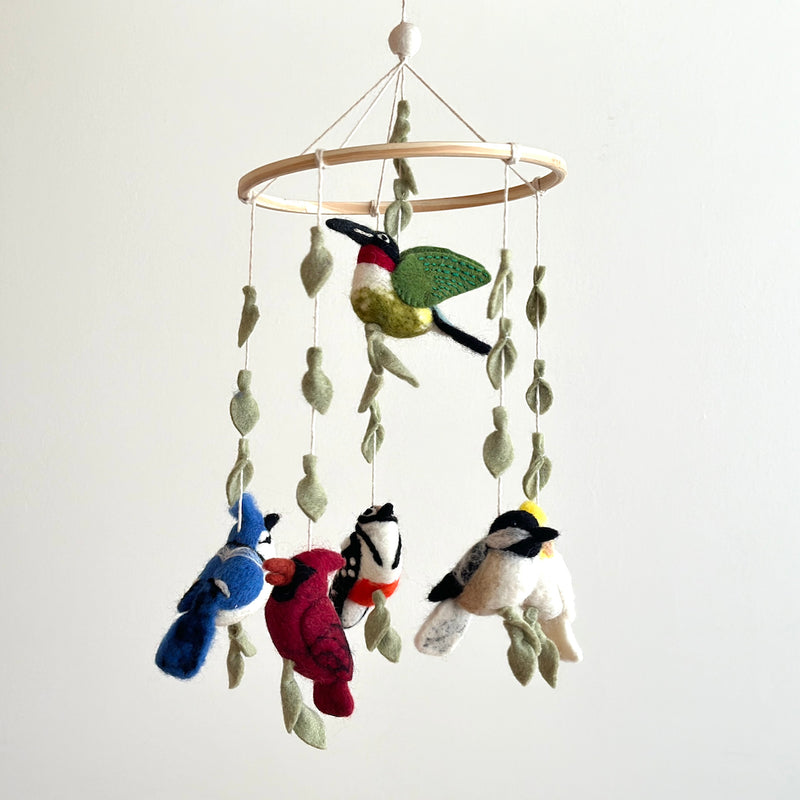 A vibrant felt bird baby mobile, titled 'Wings of America,' hangs suspended against a plain white background. Colorful felt birds and olive green felt leaves are suspended by thin white strings from a natural wooden frame. The mobile features a green, black, and red felt bird at the top, with a blue jay, cardinal, and two black and white chickadees hanging below, each accompanied by leaves.