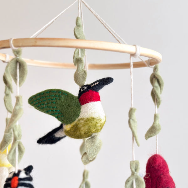 A close-up view shows a Felt Bird Baby Mobile, featuring a handcrafted wool felt hummingbird suspended from a light wooden frame. The detailed hummingbird has a black beak, red throat, white chest, and green wings with green stitching. Soft green felt leaves and other felt birds are also suspended, creating a calming, intricate design perfect for a nursery.