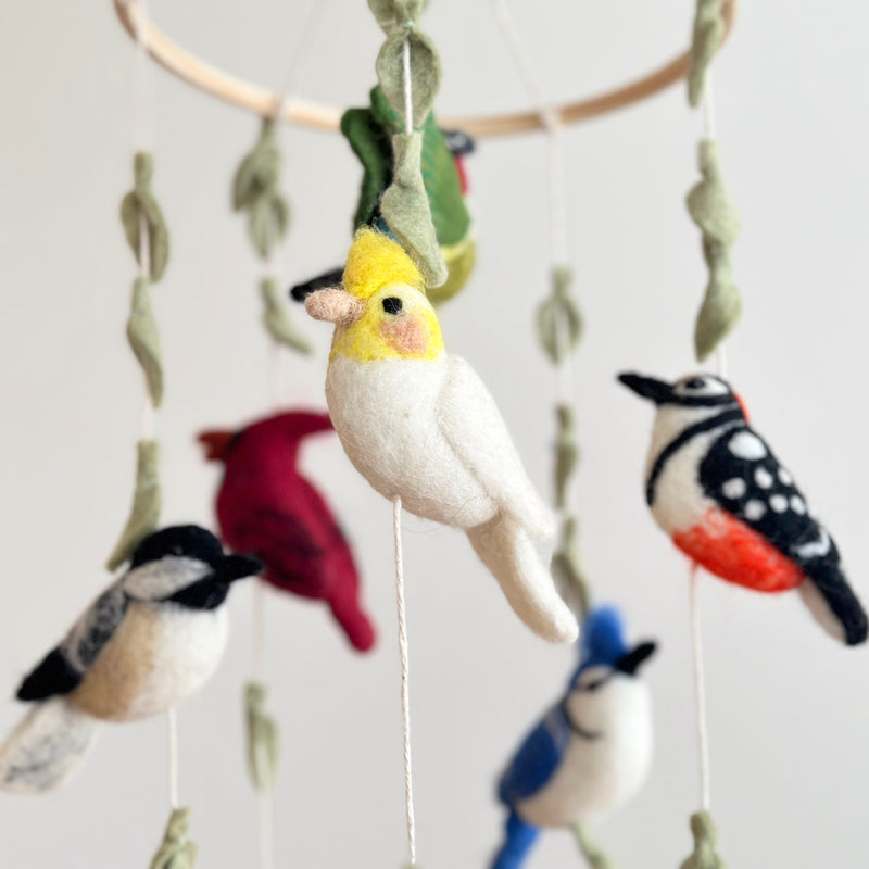 A handcrafted felt bird baby mobile, 'Wings of America,' is shown in a close-up against a plain, light-colored wall. It features a wooden frame suspending an array of colorful felt birds, including a white and yellow parrot, blue jay, red cardinal, chickadee, and green hummingbird. Green felt leaves are evenly spaced between the birds, creating a soft and inviting nursery baby mobile.