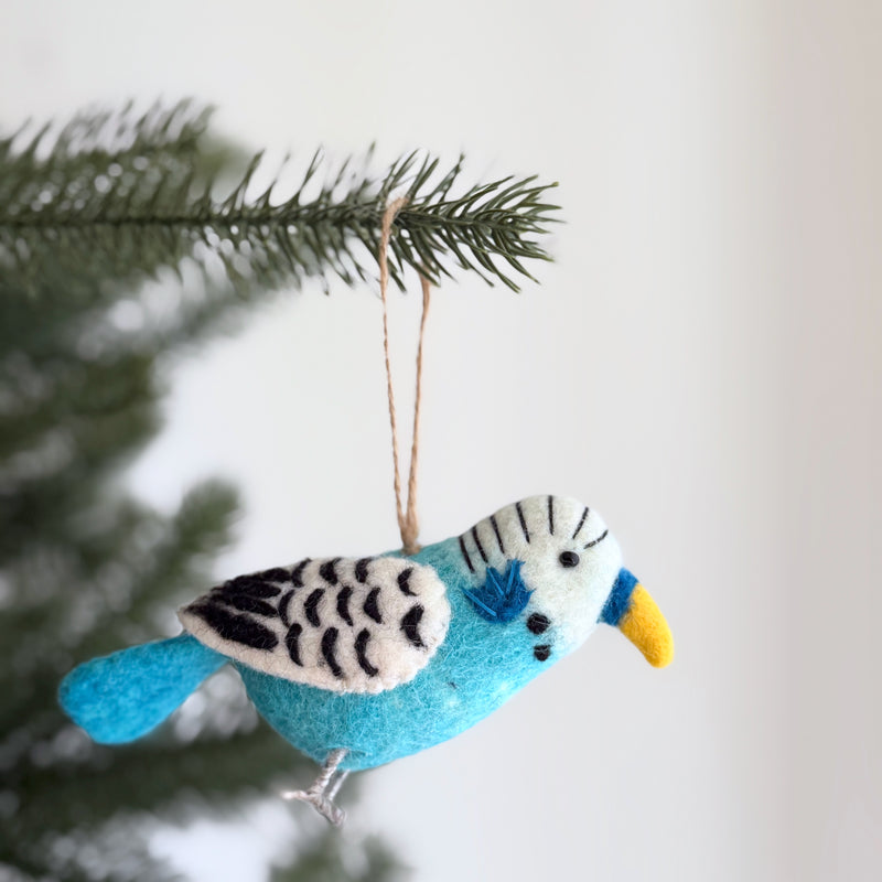 A felt blue budgerigar ornament hangs on a green Christmas tree branch against a soft white background. This cheerful wool felt Christmas ornament features a light blue body, a white head with black stripes, and black and white patterned wings. It has a yellow beak, black eyes, and delicate wire legs, complete with a jute string for hanging.