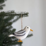 A handmade wool felt seagull ornament from the Felt Bird Ornaments set dangles gracefully from an evergreen Christmas tree branch against an off-white background. This distinctive seagull features a predominantly white body with gray striped wings, a small black eye, and bright orange beak and feet. This charming Christmas tree ornament, crafted from wool felt, adds a playful seasonal touch to holiday decor.