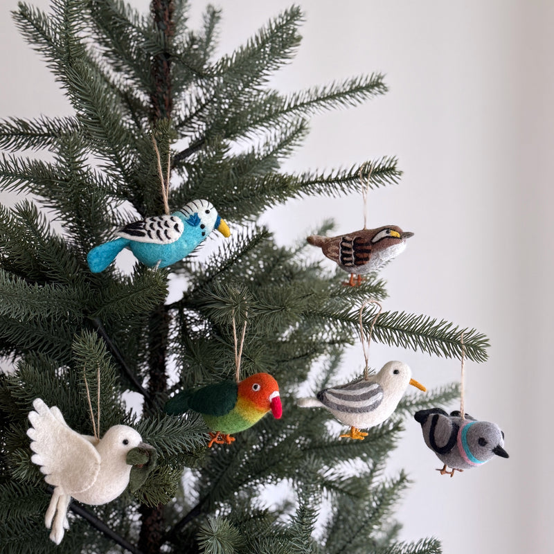 Six handmade wool felt bird ornaments, a blue budgerigar, white throat sparrow, green parrot, grey seagull, white dove, and grey pigeon, are suspended from a festive Christmas tree. Each handcrafted felt ornament features quirky details and wire legs, with the white dove holding a small green leaf. This set adds a playful, whimsical touch against a soft white backdrop.