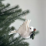 A wool felt dove ornament, part of the Felt Bird Ornaments set, hangs from a jute string against a blurred Christmas tree. The cream-colored dove, with outstretched wings and a small dark eye, gently clutches an olive branch with muted green leaves in its beak, evoking a peaceful, festive mood.