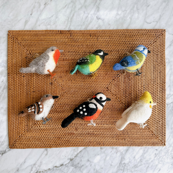 Six charming wool felt bird Christmas ornaments are arranged on a brown woven rattan placemat, which rests on a white marble surface. This set of handcrafted felt ornaments includes a robin, green canary, blue tit, sparrow, woodpecker, and white parrot, each featuring intricate needle-felted details and wire legs. Each bird boasts unique coloring, like the robin's grey and red, the canary's green, yellow, and light blue, and the woodpecker's black, white, and red.