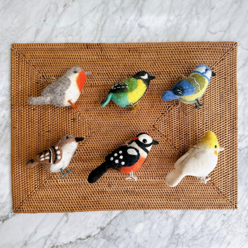 Six charming wool felt bird Christmas ornaments are arranged on a brown woven rattan placemat, which rests on a white marble surface. This set of handcrafted felt ornaments includes a robin, green canary, blue tit, sparrow, woodpecker, and white parrot, each featuring intricate needle-felted details and wire legs. Each bird boasts unique coloring, like the robin's grey and red, the canary's green, yellow, and light blue, and the woodpecker's black, white, and red.