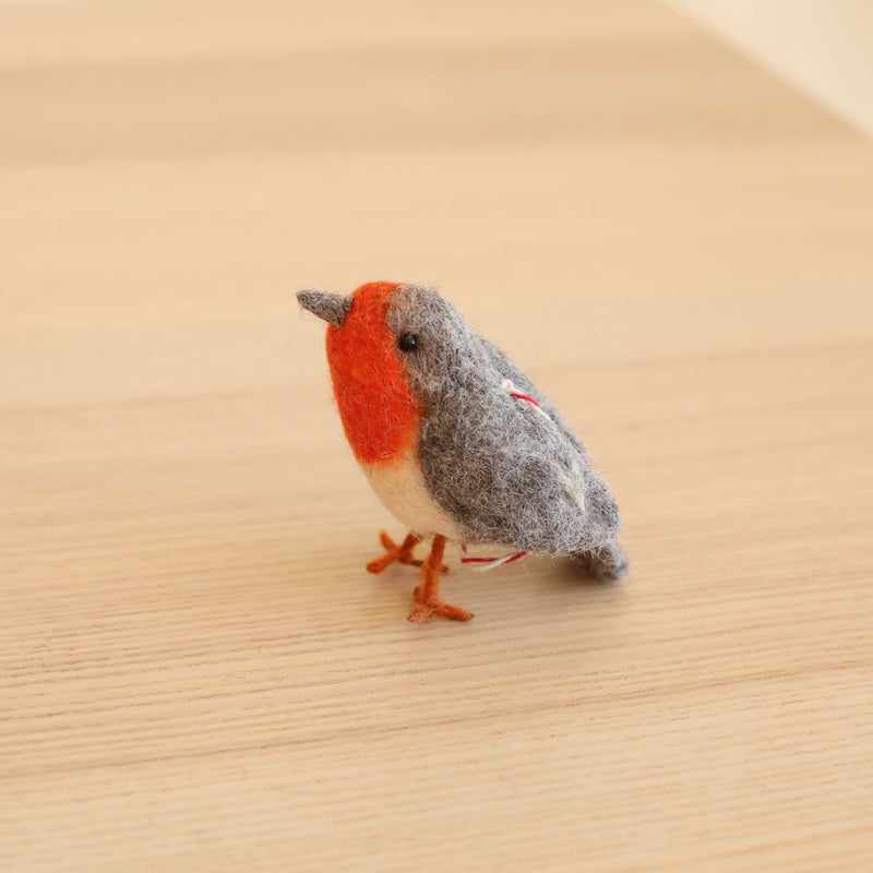 A needle-felted wool robin ornament is displayed on a light wooden surface. This handmade felt bird ornament features a gray head, a vibrant red chest, orange wire legs, and a small black eye. A red cotton string is attached to its back for hanging.
