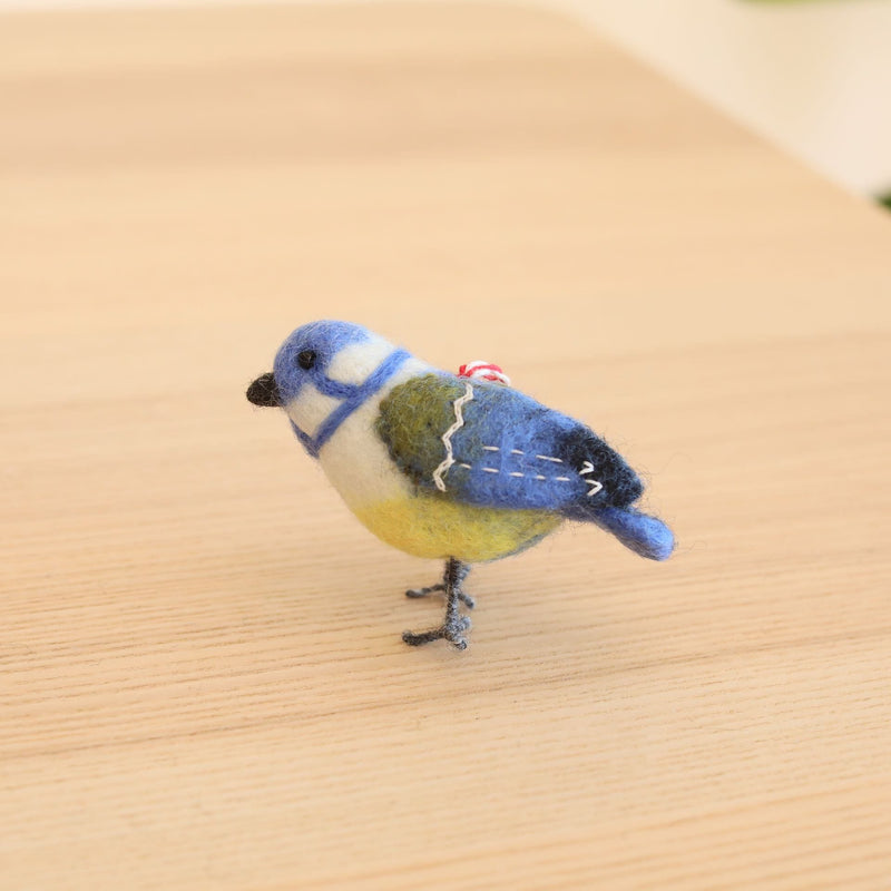 A needle felted blue tit ornament, part of a felt bird ornaments set, rests on a light wooden surface against a bright background. This detailed Christmas ornament, crafted from 100% wool, features a blue head and tail, a white and green body, and a yellow lower part. Tiny black eyes, delicate grey wire legs, and a red and white cotton string for hanging complete this charming bird.