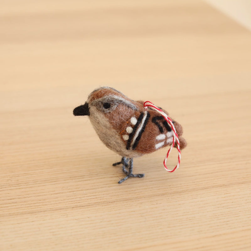 A charming needle-felted wool felt sparrow ornament, part of a set of six felt bird ornaments, rests on a light-colored wooden surface. This realistic sparrow features a plump body of brown, grey, and white wool felt, with black and white markings on its wings. It has a small black beak, delicate wire legs, and a red and white cotton hanging string.