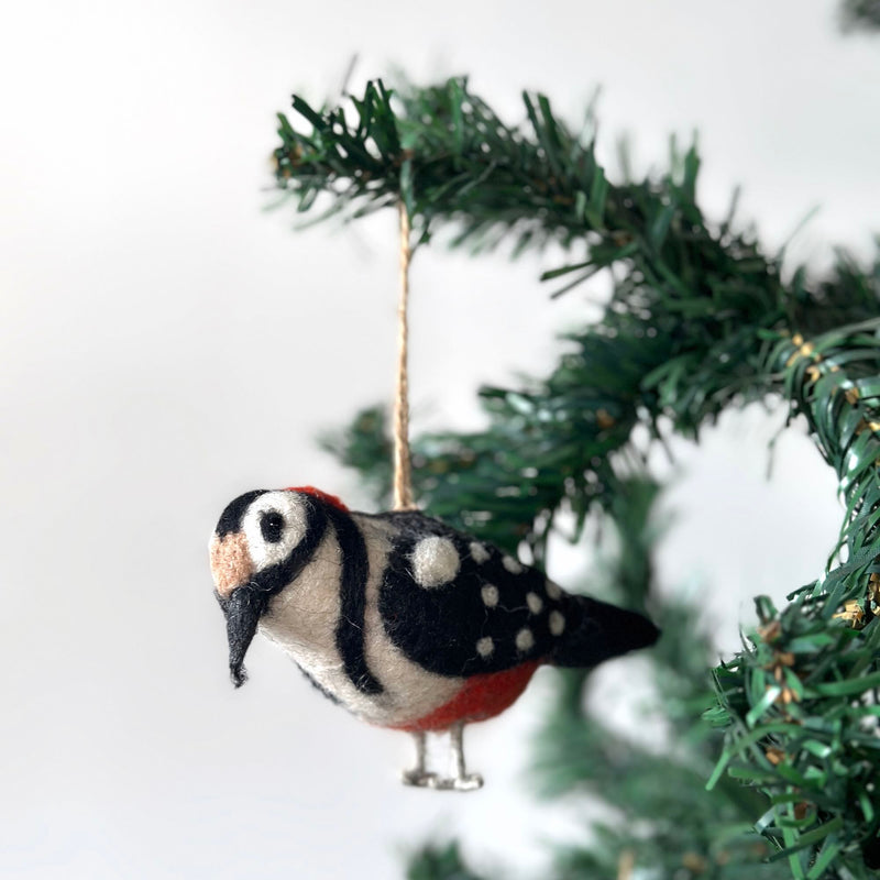 A charming wool felt woodpecker ornament, part of a festive bird ornament set, hangs gracefully from a Christmas tree branch against a clean white backdrop. This handcrafted felt woodpecker ornament features a vibrant mix of white, black, red, and tan wool with needle-felted details. Distinctive features include a red patch on its head, black stripes on its sides, and wire legs.