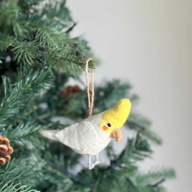 A handmade needle felted white parrot ornament, from a set of unique Christmas felt bird ornaments, is showcased hanging on a vibrant green Christmas tree. The white parrot features a predominantly white body with a bright yellow crest, a small light brown beak, and a touch of orange near its eye. It hangs gracefully by a jute string among the pine needles, next to a natural pine cone.