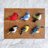 A set of six felt bird ornaments is neatly arranged on a woven mat, viewed against a bright marble background. This cheerful collection of handcrafted felt ornaments includes a cardinal, bluejay, hummingbird, chickadee, goldfinch, and kingfisher. Each unique bird features vibrant color patterns and delicate wire legs.