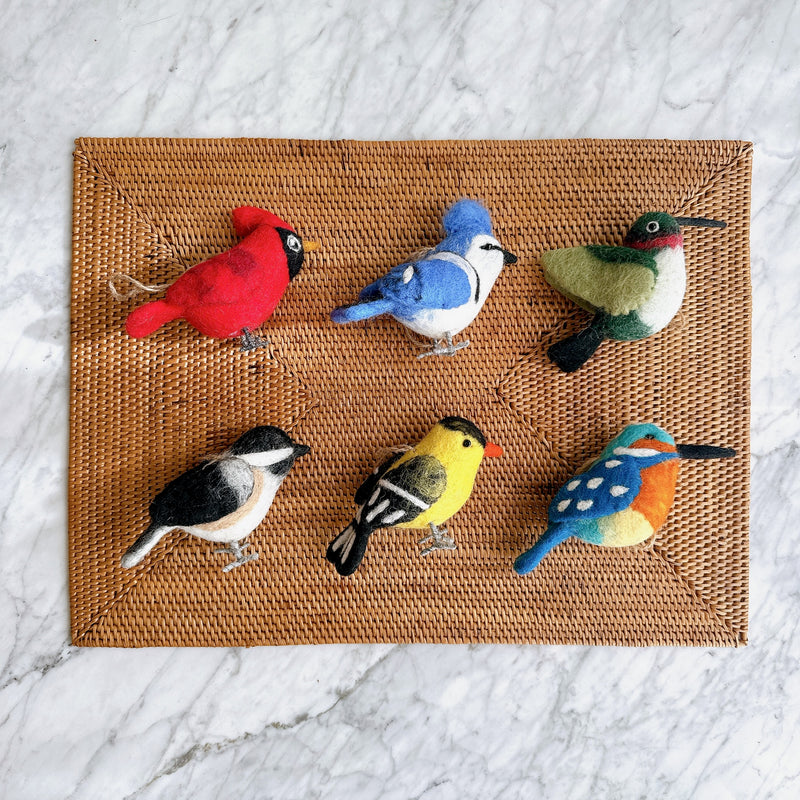 A set of six felt bird ornaments is neatly arranged on a woven mat, viewed against a bright marble background. This cheerful collection of handcrafted felt ornaments includes a cardinal, bluejay, hummingbird, chickadee, goldfinch, and kingfisher. Each unique bird features vibrant color patterns and delicate wire legs.
