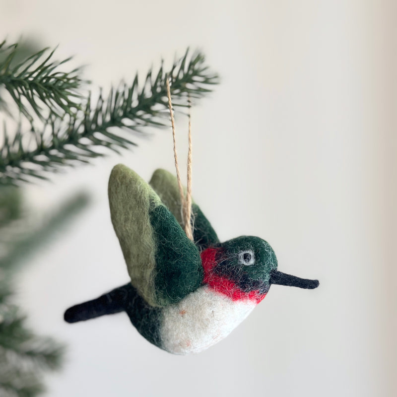 A handcrafted wool felt hummingbird ornament hangs from a deep green Christmas tree branch, set against a soft, neutral backdrop. The needle-felted hummingbird features light green wings with darker accents, a white front, and a dark green head with red markings. A slender black beak extends from its face, and a jute string is attached for easy hanging.