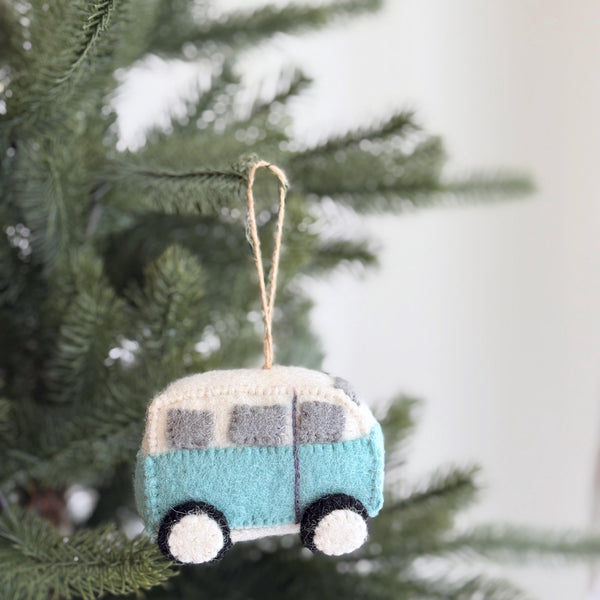 A Retro Wool Felt Blue Camper Van Christmas Ornament hangs on a Christmas tree. This handmade felt camper van ornament features a light blue body, a white roof, and gray windows. Made from 100% wool felt, it has a soft, textured appearance with round black and white wheels. It hangs ready for display from a jute string, adding a joyful, adventurous touch.