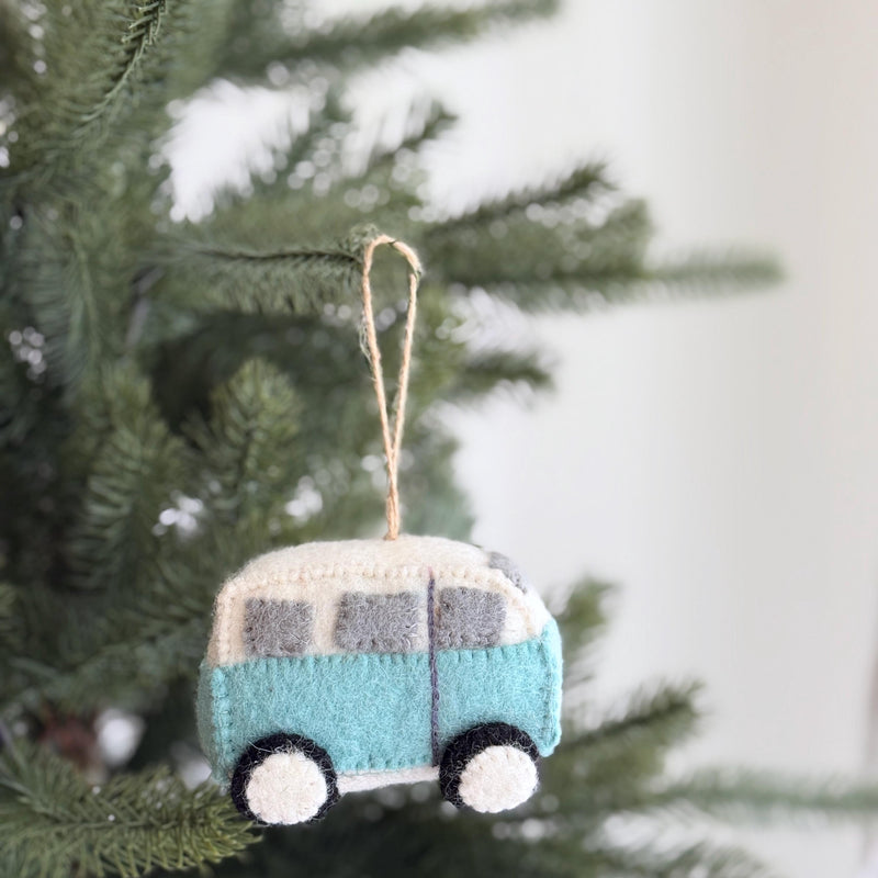 A Retro Wool Felt Blue Camper Van Christmas Ornament hangs on a Christmas tree. This handmade felt camper van ornament features a light blue body, a white roof, and gray windows. Made from 100% wool felt, it has a soft, textured appearance with round black and white wheels. It hangs ready for display from a jute string, adding a joyful, adventurous touch.