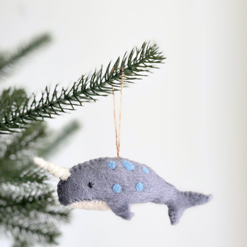 A handmade felt blue narwhal ornament is delicately hung on a Christmas tree branch, set against a bright, festive background. This unique wool felt narwhal ornament features a soft gray body with playful blue spots and a white belly. It has black eyes and a distinctive stitched ivory tusk, with a jute string attached for hanging.