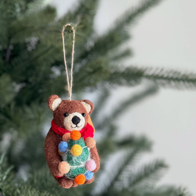 A felt brown bear Christmas ornament hangs on a green Christmas tree. Crafted from soft wool felt, the bear wears a red scarf and holds a small Christmas tree decorated with colorful lightbulbs. It features small, rounded ears and a light brown muzzle. A jute string allows for easy hanging, adding a cheerful touch to holiday decor.