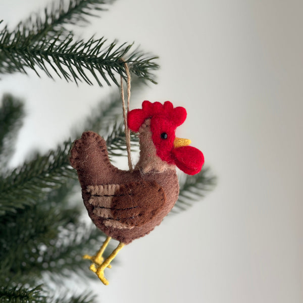 A little brown felt rooster ornament hangs from a green Christmas tree branch, presented against a neutral background. This handcrafted felt ornament features a dark brown body with subtle horizontal stripes, a red head crest and wattle, and distinctive yellow felt legs. It is suspended by a simple jute string, evoking a joyful, rustic, and handcrafted appeal.