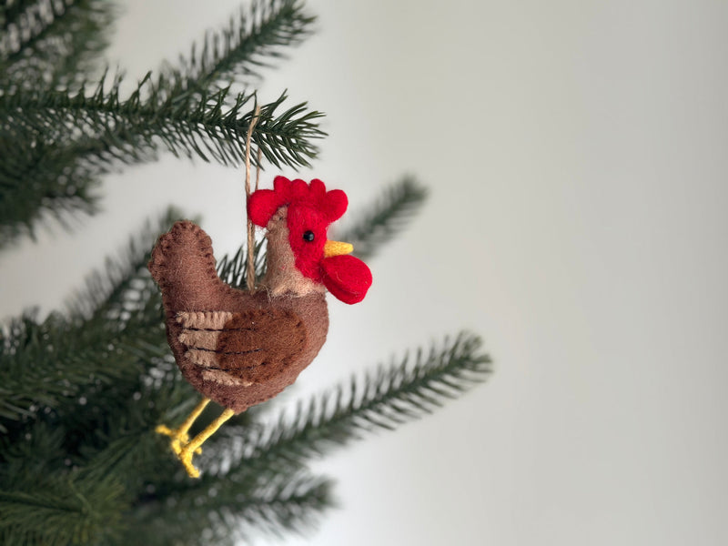 A handcrafted felt brown rooster ornament hangs suspended by a jute string on a vibrant green pine branch, nestled among its needles. This unique felt rooster ornament features a body with a gradient of brown shades and lighter horizontal stripes. Its head is bright red with a small yellow beak and tiny black eye, complemented by yellow felt feet.