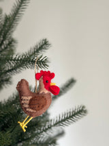 A felt brown rooster ornament hangs on the pine branches of a Christmas tree, set against a soft white background. This charming Christmas tree ornament features a brown body with tan stripes, a red head, and a yellow beak, complete with a jute string for hanging.