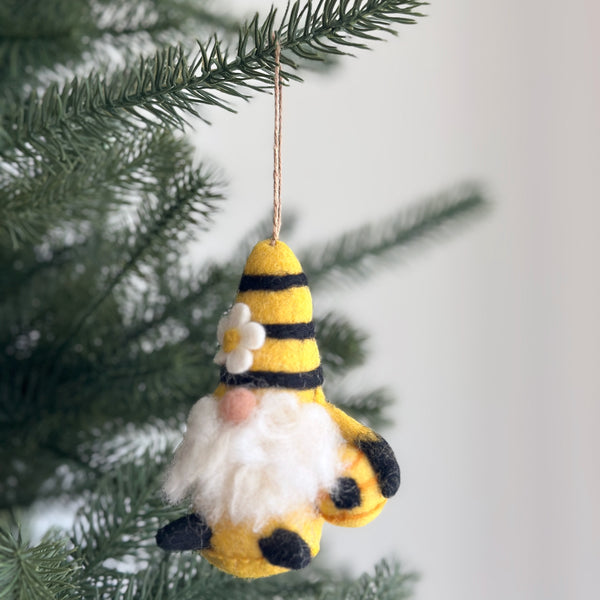 A handcrafted needle felted bumblebee gnome ornament hangs on a pine tree branch, suspended by a thin jute string, against a bright background. This unique Christmas ornament features a conical hat with yellow and black stripes, adorned with a small white flower. A fluffy white beard extends from beneath the hat, and the gnome holds a small, rounded beehive.