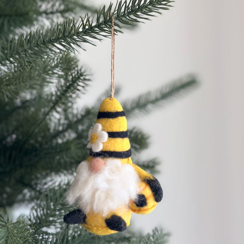 A handcrafted needle felted bumblebee gnome ornament hangs on a pine tree branch, suspended by a thin jute string, against a bright background. This unique Christmas ornament features a conical hat with yellow and black stripes, adorned with a small white flower. A fluffy white beard extends from beneath the hat, and the gnome holds a small, rounded beehive.