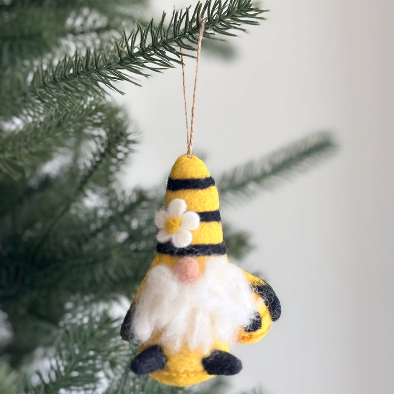 A charming wool felt bumblebee gnome ornament hangs from a green pine branch, set against a soft gray background. This needle felted gnome features a yellow striped pointed hat with a white flower, a fluffy white beard, a pink nose, and black shoes. It is holding a small beehive, and a jute string is attached for hanging.