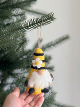 A handcrafted wool felt bumblebee gnome ornament is presented hanging from a lush green Christmas tree branch, held by a female hand. The charming felted gnome features yellow and black stripes, a fluffy white beard, and a pointed hat adorned with a white flower. A jute string loop is attached for easy hanging.