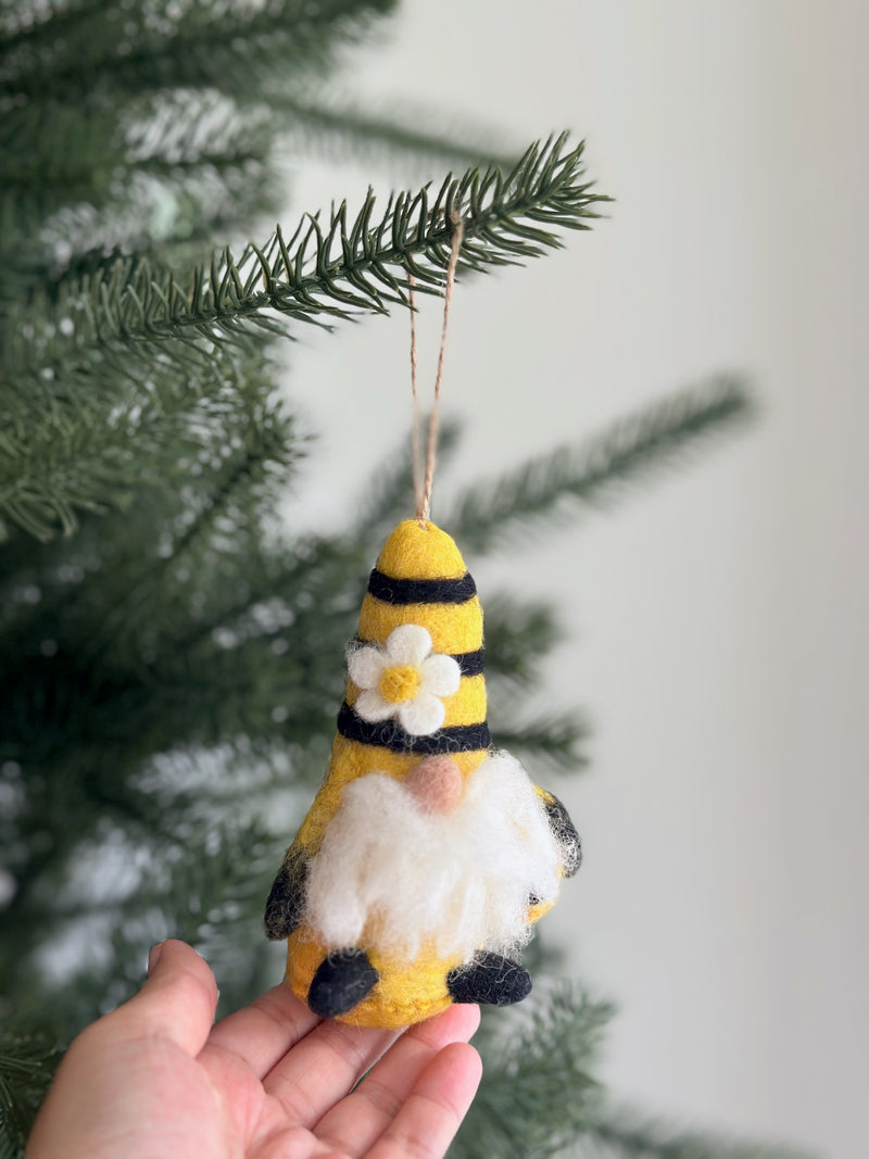 A handcrafted wool felt bumblebee gnome ornament is presented hanging from a lush green Christmas tree branch, held by a female hand. The charming felted gnome features yellow and black stripes, a fluffy white beard, and a pointed hat adorned with a white flower. A jute string loop is attached for easy hanging.