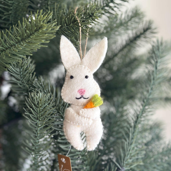 A charming white felt bunny ornament hangs against a soft, blurred green background. This handmade felt bunny ornament features long ears, black eyes, a pink nose, and a stitched black mouth. It holds a small, vibrant felt carrot with green leafy tops, and has a jute string for hanging.