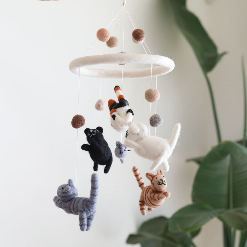 A handcrafted felt cat baby mobile is charmingly displayed against a soft white background, with a verdant plant creating a natural backdrop. Five whimsical felt kittens, in gray, black, white, tabby, orange tabby, and calico colors, are suspended from a circular white felt frame. Each adorable kitten appears to be chasing a small gray felt mouse, creating a playful and comforting scene.