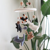 A handcrafted wool felt cat baby mobile is suspended against a softly lit white wall, with lush green plant leaves subtly blurred in the background. This charming felt baby mobile features five adorable felt kittens in various colors like white, black, gray, and different tabby patterns, playfully chasing a small gray felt mouse. All characters display intricate hand-stitched details.
