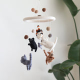A felt cat baby mobile hangs against a clean, bright white wall, softly lit with a glimpse of greenery. This handcrafted felt baby mobile features five adorable, hand-stitched felt kittens in various colors, surrounding a smaller gray mouse. The central ring is covered in soft white felt, adding a gentle accent.