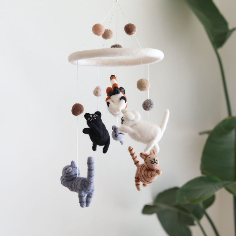 A felt cat baby mobile hangs against a clean, bright white wall, softly lit with a glimpse of greenery. This handcrafted felt baby mobile features five adorable, hand-stitched felt kittens in various colors, surrounding a smaller gray mouse. The central ring is covered in soft white felt, adding a gentle accent.