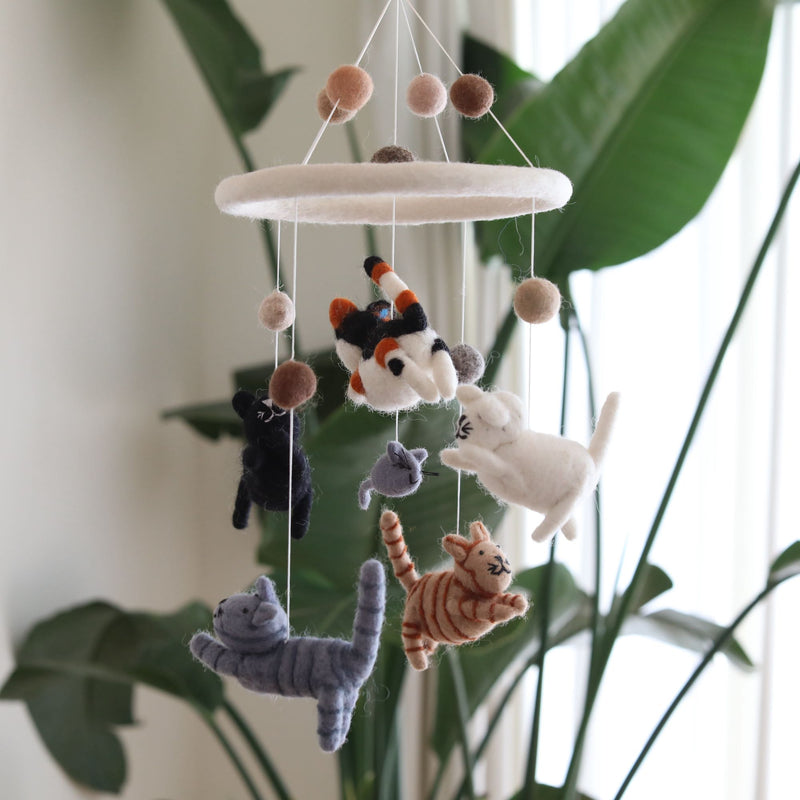 A playful Felt Cat Baby Mobile hangs in a softly lit, natural setting, with a blurred green houseplant and sheer white curtains in the background. Six adorable felt kittens—white, black, gray, tabby, orange tabby, and calico—along with a small gray felt mouse, dangle from a circular white felt ring on thin strings. Small brown fuzzy balls also adorn the strings, creating a comforting and visually engaging nursery accessory.