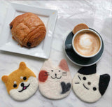 Three round, handmade wool felt cat and dog coasters, part of a Felt Cat & Dog Coaster set, are displayed on a marble table alongside a coffee cup and pastry. These ethically crafted coasters include a white and orange shiba inu, a white cat with a black heart, and a white and black tuxedo cat design. These charming, needle felted creations by Nepalese female artisans add a whimsical touch to the cozy scene.