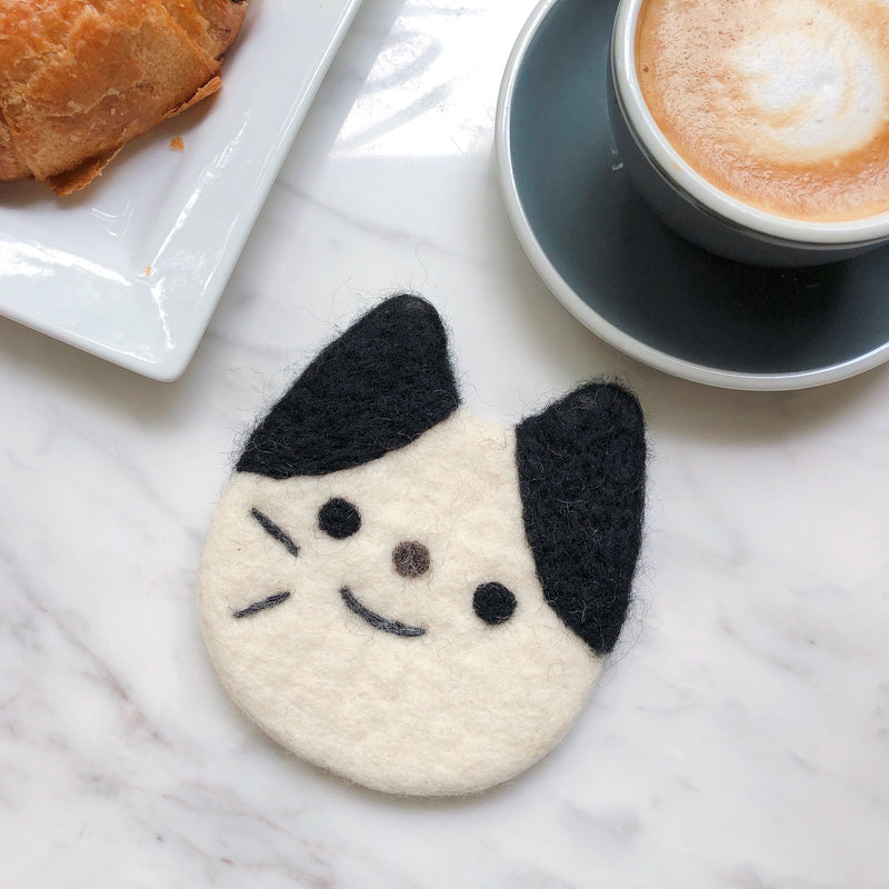 A white wool felt cat coaster, ethically handcrafted by Nepalese female artisans, rests on a marble table with a coffee cup and croissant. This charming cat coaster, part of a set, is round with black ears and facial markings, featuring stitched details for a cute and playful expression, adding a whimsical touch to the cozy scene.