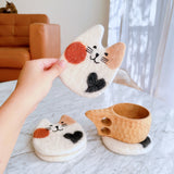 These handcrafted needle felted calico cat coasters are presented on a bright marble table, with one held by a hand and another supporting a wooden mug, creating a cozy and playful scene. Each unique, environmentally friendly coaster is made from ethically sourced sheep wool, featuring a charming calico cat face with a white base, an orange spot, black whiskers, perky ears, and a black heart.