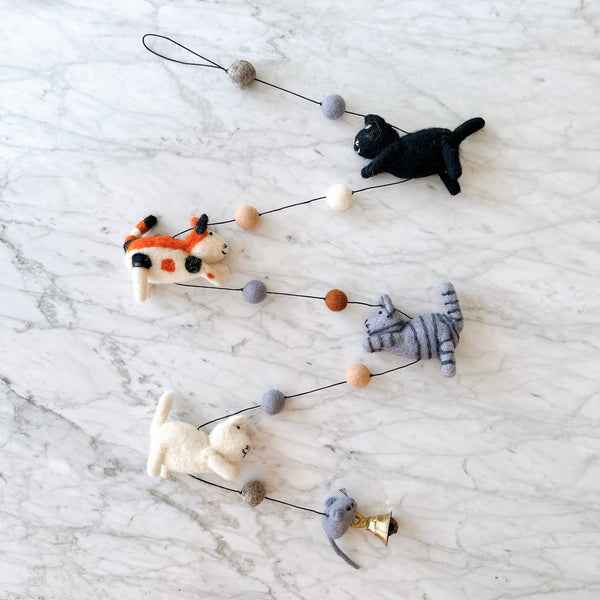 A handmade wool felt cat garland is displayed on a white marble surface. It features four felted cats in black, white, grey, and orange, strung together with small felt balls in muted earth tones. A copper bell hangs at the end of the garland.